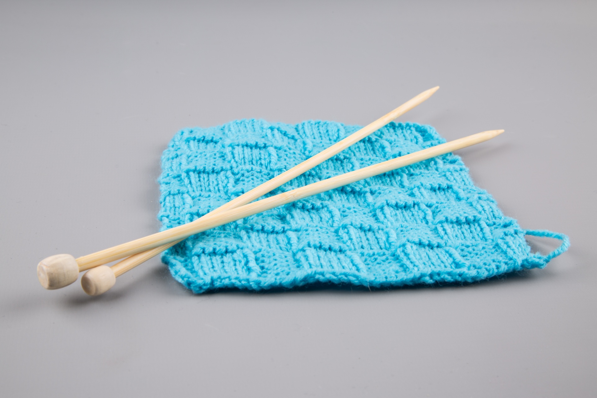 Image of a bright blue-teal knit square knit with the basket stitch.  Two wooden knitting needles rest on top of the knit square.