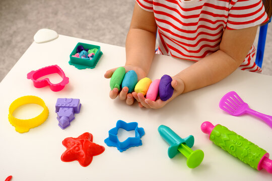 a light skinned child in a red and white striped shirt holds playdough in their hand and is surrounded by a variety of tools including molds, cookie cutters, and rolling pins