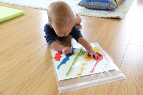 A baby playing with vibrant paint on the floor, creating a colorful artwork contained in a plastic bag.