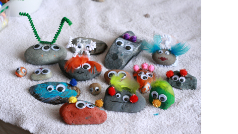 An assortment of painted rocks with googly eyes and pompoms glued on them. Some also have feathers, hair, or pipe cleaners. They are on a gray background facing the camera.
