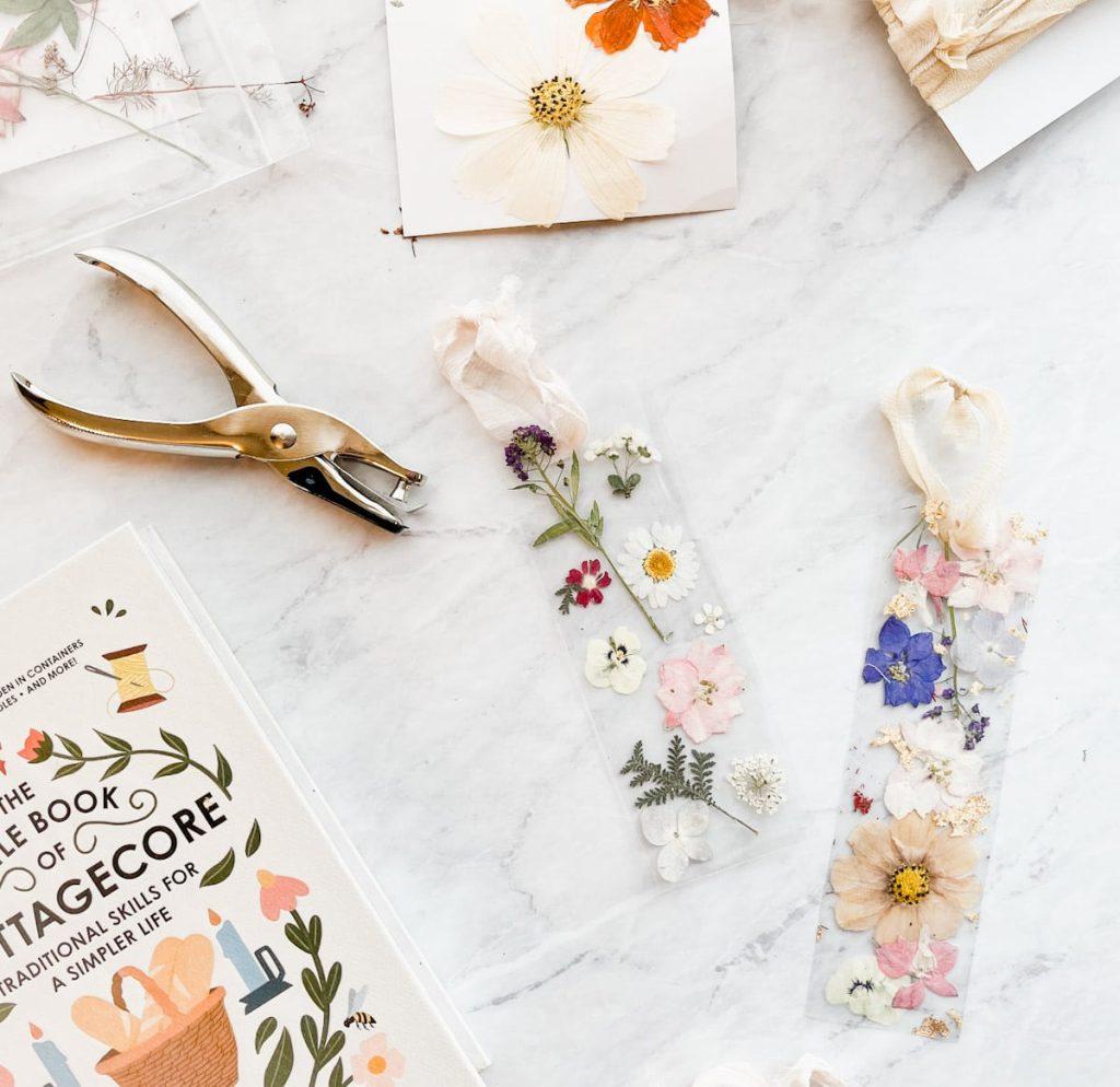 hole punch and two pressed flower bookmarks with ribbon on a marble counter next to a book